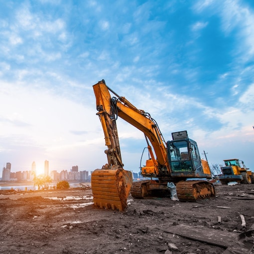Large yellow excavator parked on a muddy construction site beside a body of water, with a city skyline of modern high-rise buildings in the background.