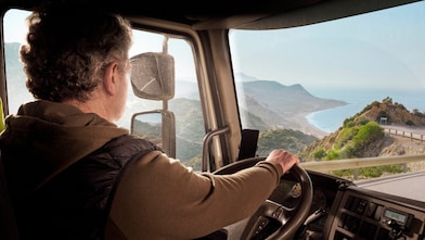 A truck driver steering along a coastal road with a scenic view of mountains and the sea.