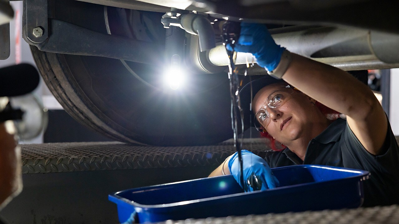 A mechanic drains oil from a vehicle into a blue container in a workshop