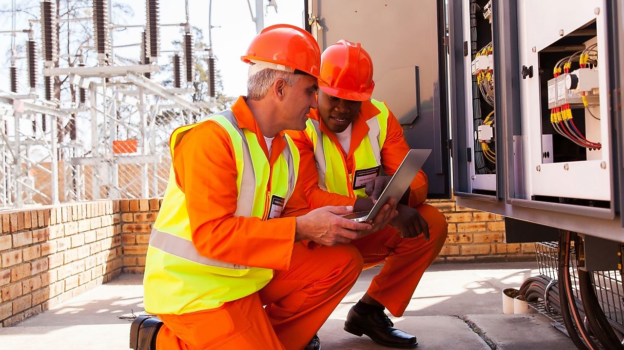 Two men wearing construction orange gaze at a laptop.