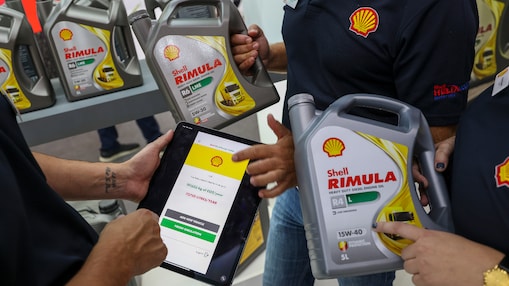 Three people holding Shell Rimula bottles and a tablet displaying Shell CO2 and Energy Tracker. The background shows shelves with more Shell Rimula bottles.