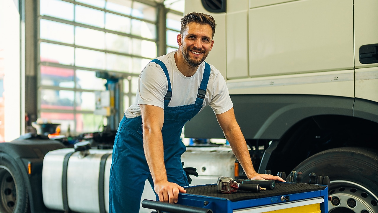 Portrait of positive smiling truck serviceman with tools standing by truck vehicle in workshop. Truck vehicle maintenance and servicing.
