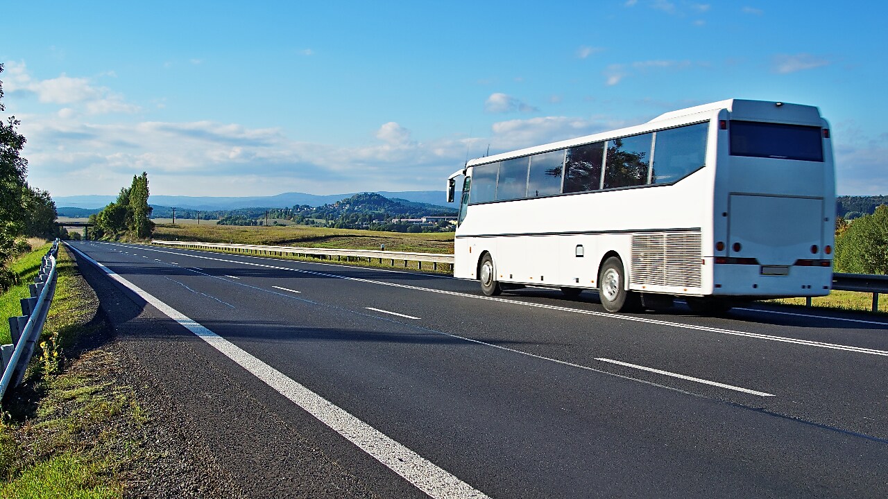 White Bus driving on an empty road in a rural landscape. Villages and forested mountains in the background.