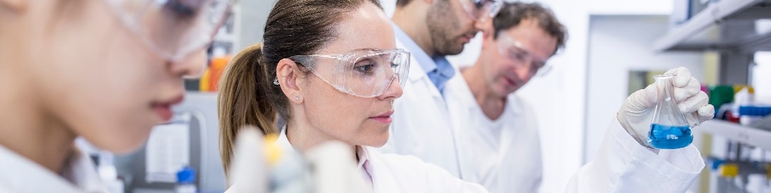 Scientists in a lab with white lab coats and safety glasses handle samples