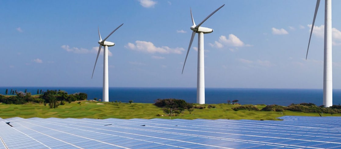 Solar panels in the foreground and three wind turbines on a coastal landscape overlooking the ocean under a blue sky with scattered clouds. The image illustrates renewable energy generation from both solar and wind power.