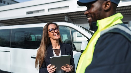 Two people conversing beside a white van, with one person holding a tablet device.