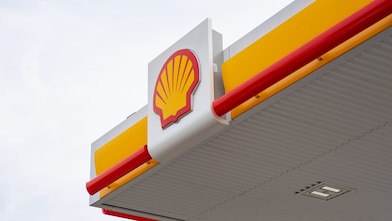Close-up of Shell petrol station canopy corner with yellow fascia, red trim, and Shell logo mounted on a white panel against a grey sky.
