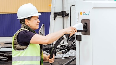 A worker in a hard hat and high-visibility vest plugging a charging connector into a Shell Recharge unit.