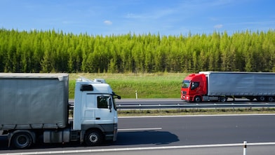 Two articulated lorries travelling in opposite directions on a divided highway bordered by trees under a clear blue sky.