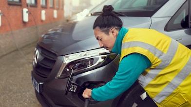 Person in high-visibility vest plugging charging cable into front of grey van, with wall-mounted chargers visible along brick building.