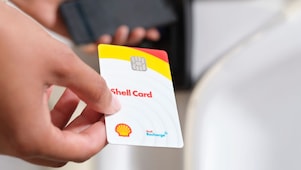 Close-up of a hand holding a Shell Card in front of a payment terminal, with another person holding a smartphone in the background.
