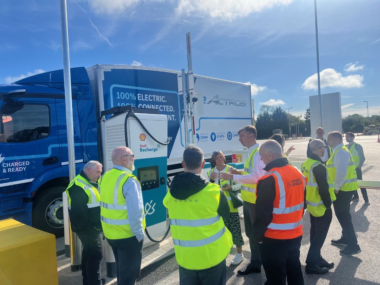 People standing in a circle next to a truck and charge point in a parking bay.