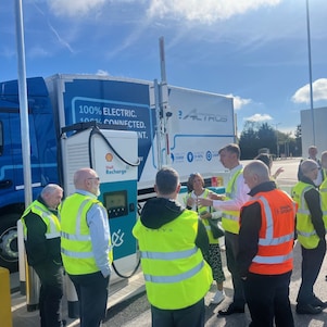 People standing in a circle next to a truck and charge point in a parking bay.