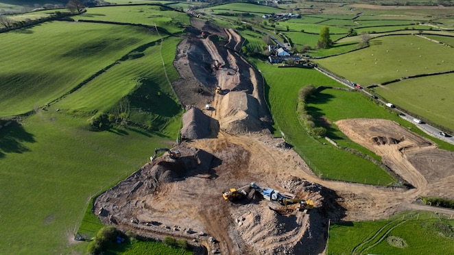 Aerial view of heavy machinery reshaping land for infrastructure development in green fields near a village