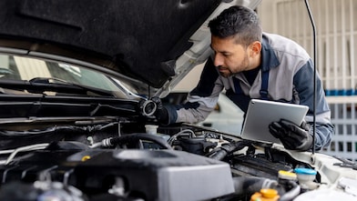Mechanic wearing gloves inspecting car engine under open bonnet while holding a tablet device.