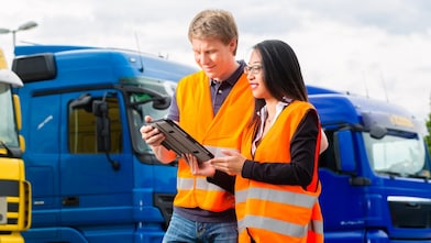 Two people wearing orange high-visibility vests reviewing information on a tablet in front of parked blue trucks.