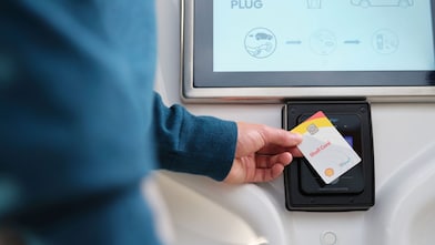 Close-up of a hand tapping a Shell Card against a contactless payment reader mounted below a screen.