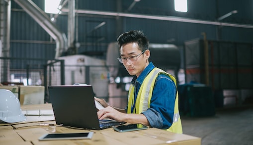 Person wearing high-visibility vest working on a laptop at a workstation in an industrial environment.