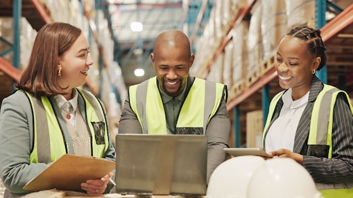 Three warehouse workers in high-visibility vests gathered around a laptop, holding clipboards and a hard hat.