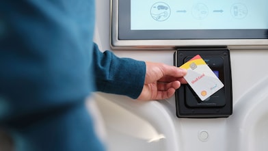 Close-up of a hand tapping a Shell Card against a contactless payment reader mounted below a screen.