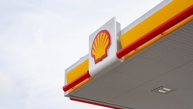 Close-up of Shell petrol station canopy corner with yellow fascia, red trim, and Shell logo mounted on a white panel against a grey sky.