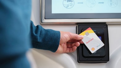 Close-up of a hand tapping a Shell Card against a contactless payment reader mounted below a screen.