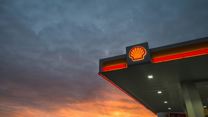 The corner of a Shell service station is against a dramatic backdrop of a cloudy sky at sunset, with an orange glow.