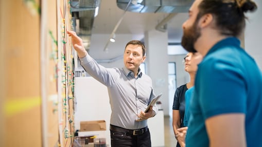 Three colleagues standing indoors reviewing a wall-mounted planning board, with one person pointing while holding a tablet.