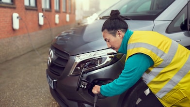 Person in high-visibility vest plugging charging cable into front of grey van, with wall-mounted chargers visible along brick building.