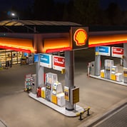 Shell petrol station forecourt at night, with illuminated canopy, Shell logo and fuel pump islands labelled AutoGas, V-Power and AdBlue.