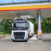 Volvo lorry refuelling under a Shell canopy at pumps labelled AdBlue 13 and 14, with a display stand of vehicle fluids nearby.