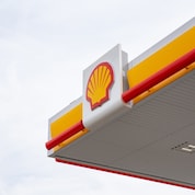 Close-up of a Shell petrol station canopy corner showing the Shell logo on a white panel with yellow fascia and red trim against a grey sky.