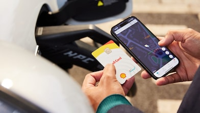 Close-up of person holding Shell Card and smartphone displaying map interface beside electric vehicle charging unit.