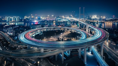 Aerial night view of circular highway interchange with long-exposure light trails and illuminated city skyline in background.