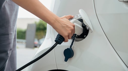 Close-up of hand inserting white electric vehicle charging connector into charging port of white car.