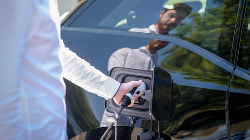 Person plugging charging connector into side charging port of black electric car, with reflection visible in car door.