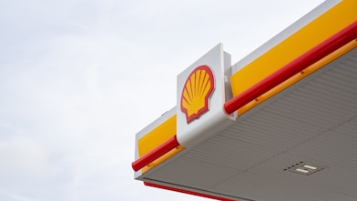 Close-up of Shell petrol station canopy corner with yellow fascia, red trim, and Shell logo mounted on a white panel against a grey sky.
