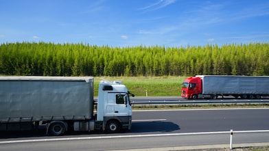 Two articulated lorries travelling in opposite directions on a divided highway bordered by trees under a clear blue sky.