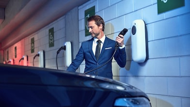 A person in suit holding electric vehicle charging connector beside car, with multiple wall-mounted chargers in background.