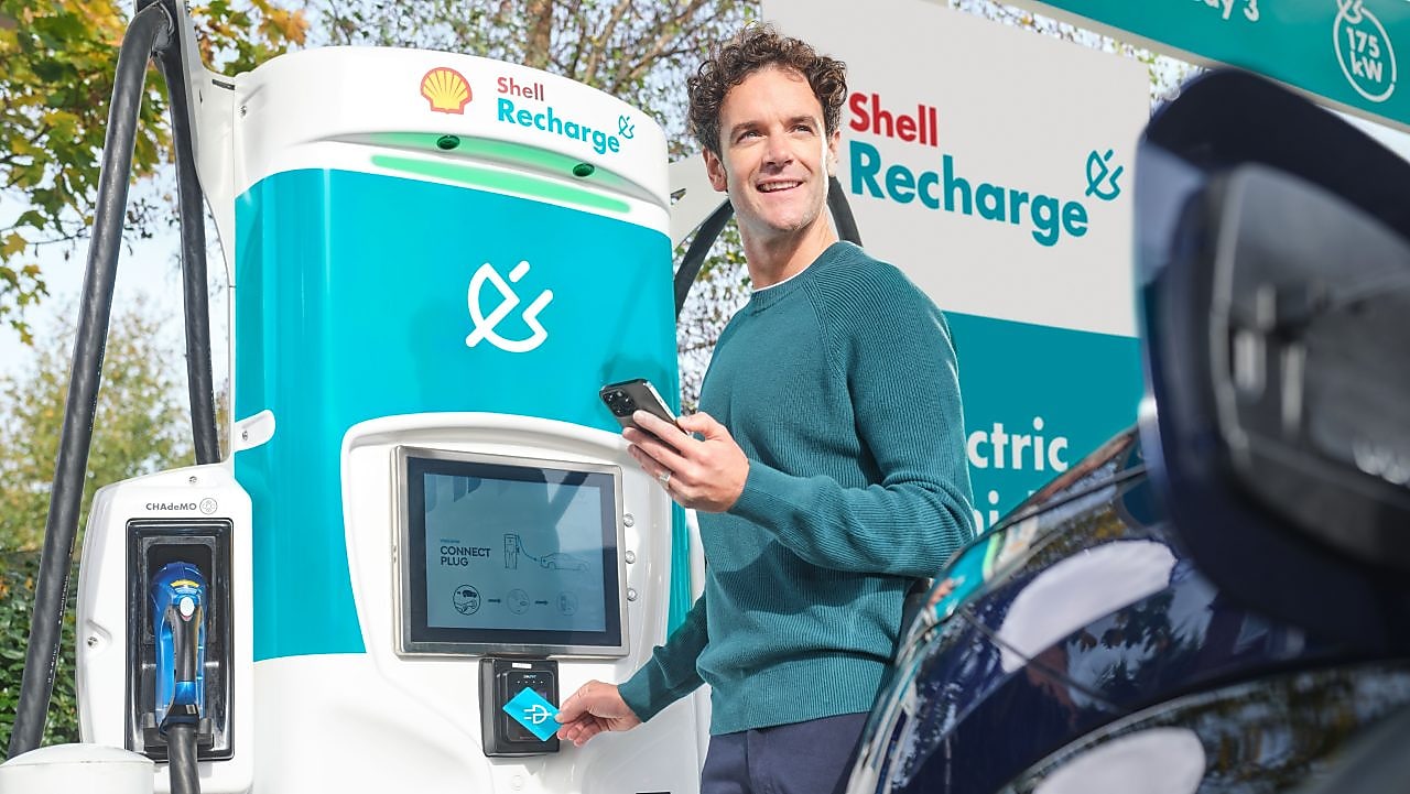 A man using a Shell Recharge electric vehicle charging station, holding a smartphone and tapping his charge card on the charger