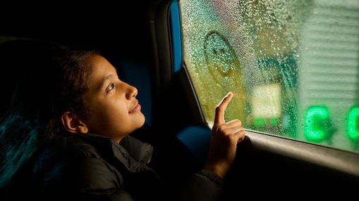 A child sitting inside a vehicle traces a smiley face on a misted side window with a fingertip. Outside the window, the green indicator lights of a Shell Recharge electric vehicle charge point glow softly in the background. The image is lit by low, warm light, with water droplets on the glass creating a textured effect and highlighting a moment during an EV charging stop.