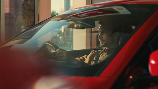 The image shows a close up of a man in the driving seat of a red vehicle, holding the steering wheel. A reflection at the top of the windscreen shows a Shell V-Power sign, and to the left of the car, there is a partial view of a digital forecourt screen, showing the Shell Pecten (logo).