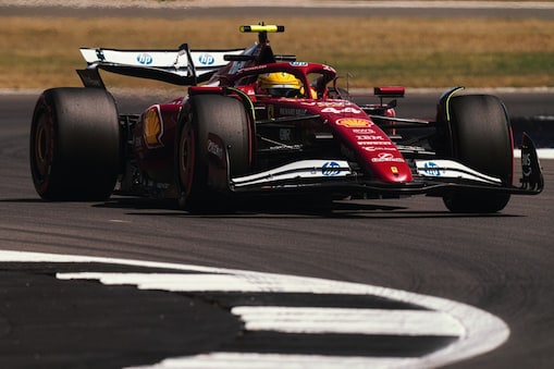 A red Scuderia Ferrari HP Formula 1 race car with prominent Shell and Ferrari logos is captured mid-turn on a racetrack. The car has large tires, aerodynamic wings, and a driver wearing a yellow helmet. The background shows part of the track and grass.