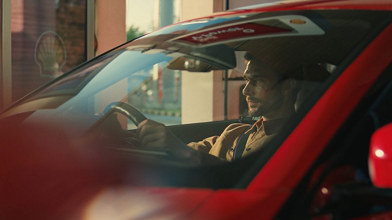 The image shows a close up of a man in the driving seat of a red vehicle, holding the steering wheel. A reflection at the top of the windscreen shows a Shell V-Power sign, and to the left of the car, there is a partial view of a digital forecourt screen, showing the Shell Pecten (logo).