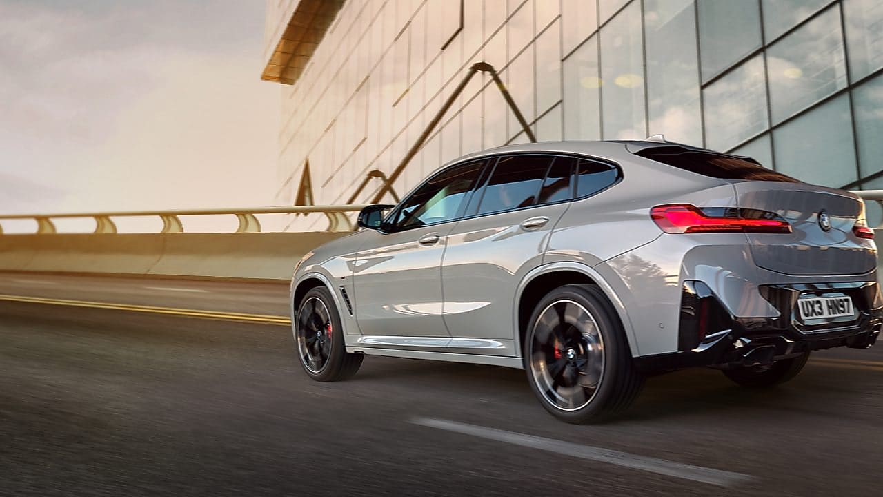 A silver BMW X4 is driving on a road next to a modern glass building with geometric patterns. The vehicle is shown from the rear and side, and the sky appears cloudy with a hint of sunlight breaking through.