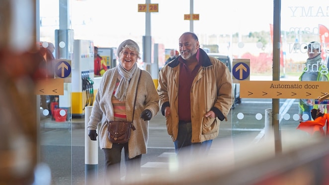 Image shows a lady and man smiling while entering a Shell service station through automatic doors. The image backdrop is a Shell forecourt.