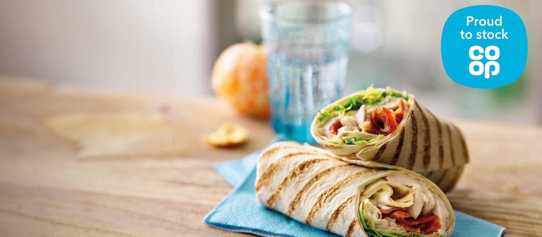 Two grilled wrap halves filled with sliced chicken, cheese, and salad leaves, stacked on a blue napkin on a wooden table, with a blurred drink in the background. A blue Proud to Stock Co-op logo appears in the top right corner.