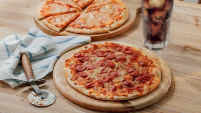 A whole pizza topped with melted cheese and sliced pepperoni sits on a wooden board on a table. A pizza cutter and tea towel are beside it, with a second pizza partially visible in the background and a tall glass of fizzy drink.