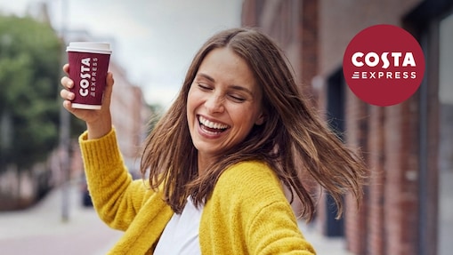 Person holding a Costa Express takeaway coffee cup while moving along a city street, wearing a mustard yellow jumper, with brick buildings in the background. The Costa Express logo appears in the top right corner.