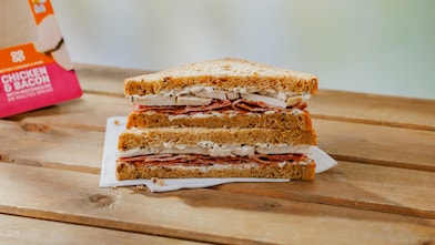 A stacked sandwich made with brown bread, sliced chicken, bacon and mayo sits on a wooden table with a napkin underneath.